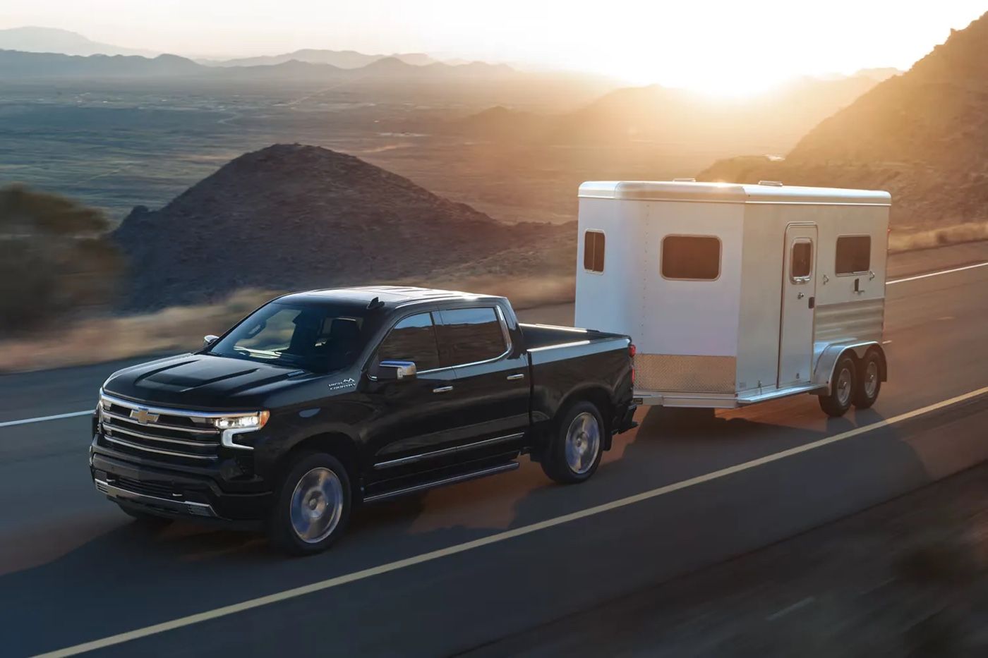 Camion Chevrolet Silverado 1500 2026 noir tractant une remorque sur route de montagne au coucher du soleil.